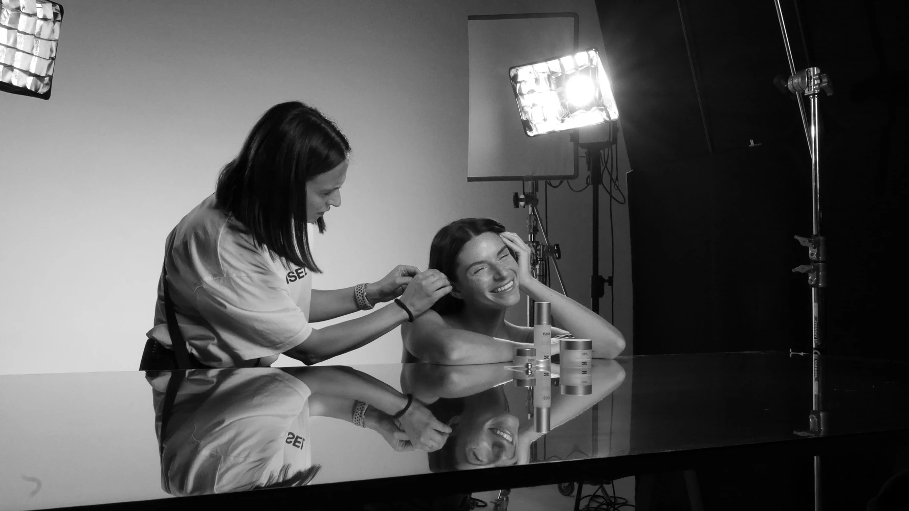 Model laughing as her hair is styled at a glossy table with skincare products